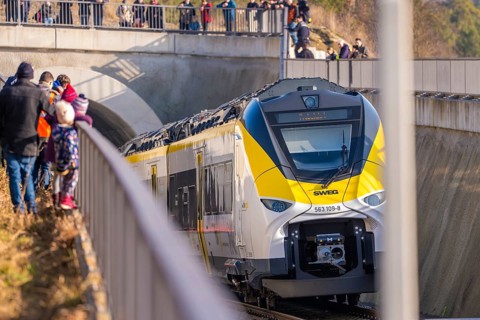 The yellow and white train passes a bridge. People are standing on the platform. Security forces