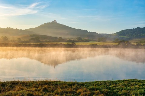 Wachsenburg Castle is reflected in the still water of a lake in the morning light, fog rises,