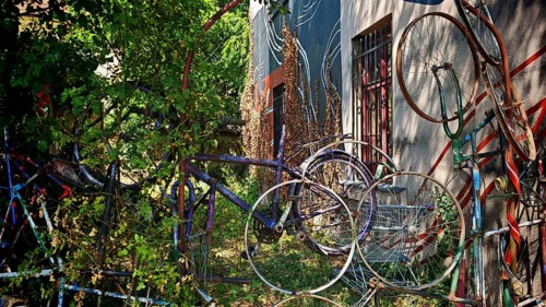 Colourful bicycles stacked on top of each other next to an art-painted wall surrounded by plants,
