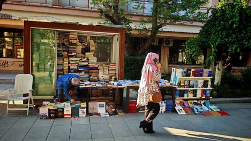 Book stand on a street surrounded by people and green trees, idyllic street scene with woman