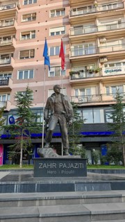 Statue of Zahir Pajaziti in front of a modern building with two flags in the background, Pristina,