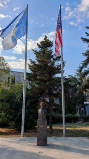 Bust statue of Madeleine Albright outdoors with two USA and Kosovo flags surrounded by tall trees,