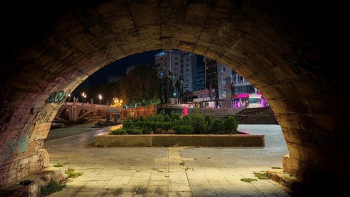 View through a stone archway of an illuminated square with monuments at night, Skopje, North