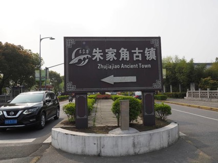 A road sign points to the ancient city of Zhujiajiao surrounded by trees and a road with cars,