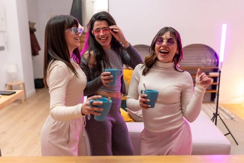 Three cheerful young female friends wearing sunglasses, laughing and holding hot drinks in mugs,
