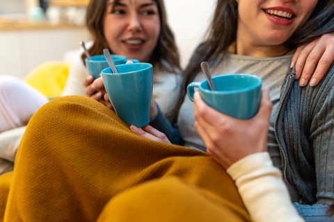 Two young women friends enjoying hot drinks and conversation while relaxing on a sofa at home,