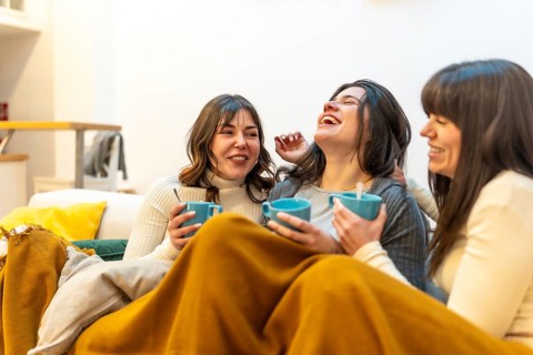 Happy women friends enjoying a cozy moment together at home, laughing and having hot drinks while