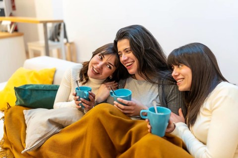 Three young women on a sofa wrapped in a blanket, sharing hot drinks, laughing and chatting in a