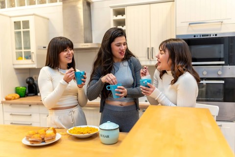 Three young women laughing and chatting around a kitchen island, sharing muffins and hot drinks in