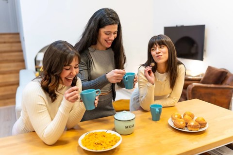 Three young women smiling and laughing, holding cups and spoons, gathering around a wooden kitchen