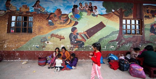 Mayan families in traditional clothes squat in front of a mural at the market in Chichicastenango,