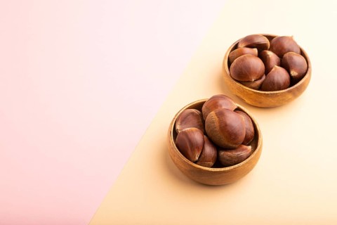 Wooden bowl with raw edible ?hestnuts on pink and orange paper pastel background, side view, copy
