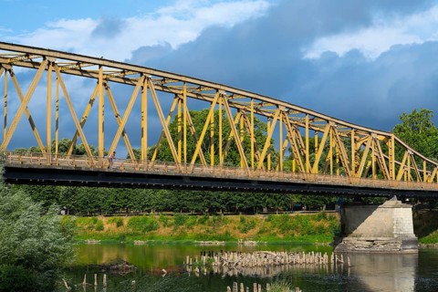 Yellow metal bridge over a river with green vegetation on the banks and cloudy sky, bridge over the