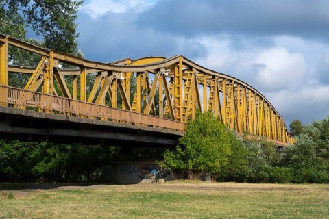 Yellow metal bridge over a meadow with trees, a river and cloudy sky in the background, bridge over