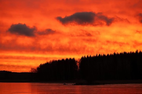 A dramatic sunset over a still, frozen lake and forest with orange-red hues in a cloudy sky,