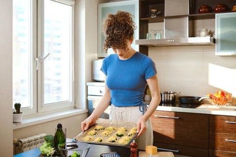 A person is in a kitchen holding a tray of smashed broccoli with cheese. Green broccoli is on top