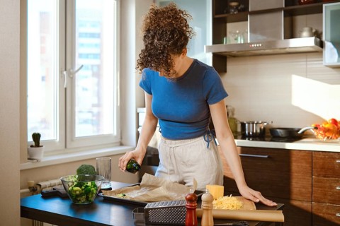 Woman pouring olive oil over the parchment. Ingredients are spread out on a counter near cooking
