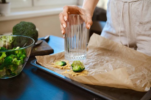 Woman is preparing smashed broccoli with cheese in a kitchen. She is using a glass to smash the