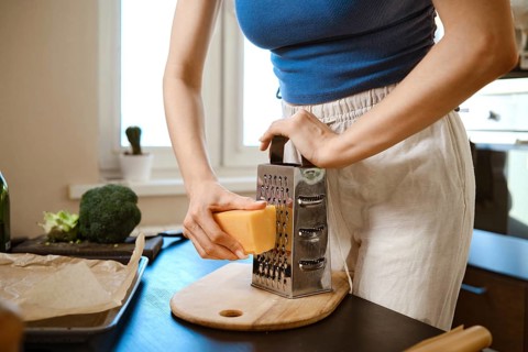 A woman grates cheese over a cutting board while preparing smashed broccoli in a kitchen. The