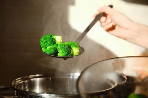 A person holds a slotted spoon with steamed broccoli above a pot in a kitchen. The steam rises,