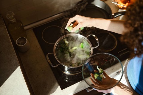 Fresh broccoli is placed into a pot of boiling water on the stove. A woman stands over the pot,