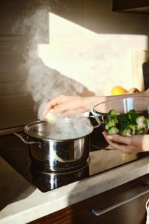 A person is cooking broccoli in a pot on the stove. Steam rises from the pot while another bowl