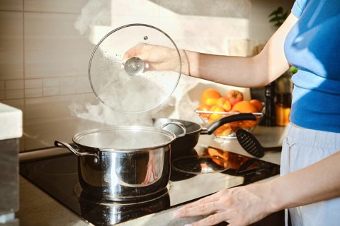 A woman lifts the lid of a saucepan in which water is boiling. Hot steam rises from the pan and is