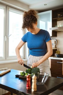 Woman chops broccoli in a kitchen. The cutting board is set on a table beside a bowl. Seasoning