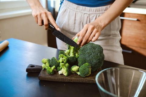 Close-up view of cutting fresh broccoli on a wooden board in a home kitchen. The person uses a