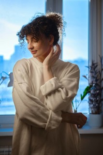 A woman stands softly with her hand on her neck in front of a window. Sunlight fills the room and