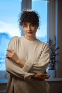 A young woman stands near a window in a dim room. She is wearing a light sweater and looks at the