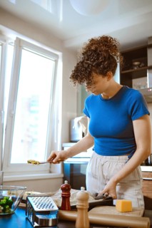 In a bright kitchen, a woman prepares smashed broccoli mixed with cheese. She stands at the