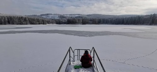 Person sitting on snowy footbridge with a view of the partially frozen lake, Rennsteig, Thuringian