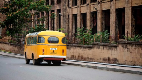 Old yellow bus runs on empty city street next to a weathered building, Alaverdi, Armenia