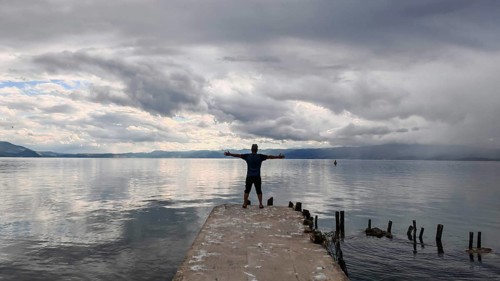 Person standing with outstretched arms on a pier surrounded by water and clouds, Lake Ohrid, North