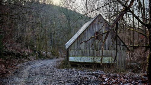 Snowy trail leads to a secluded wooden hut in the winter forest in front of a range of hills,