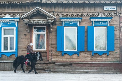Irkutsk, Siberia, Russia. A young woman riding a pony in front of traditional wooden houses during