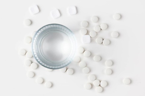 Glass of water with lots of scattered white tablets on a white surface