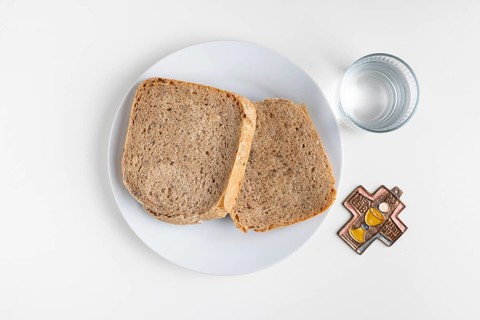 Two slices of bread on a plate with water glass and cross symbol on a plain white background, Lent