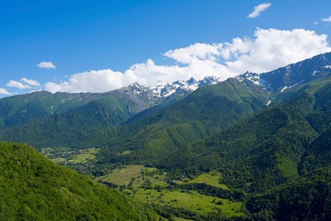 Green valley with densely wooded slopes and distant mountains under blue sky with clouds, landscape