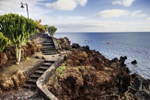 A rocky coastal path with a staircase leads to the sea under a cloudy sky