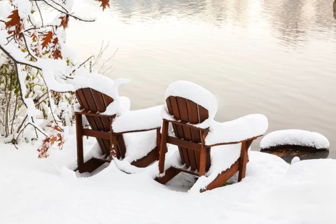 Two brown stained wooden Adirondack chairs by riverside covered with winter like snow in late