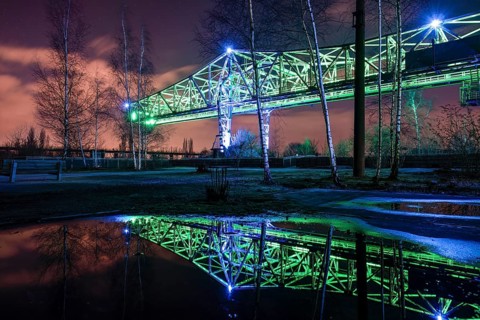 Green-lit bridge structure at night reflecting in a water surface, Landschaftspark Duisburg Nord,