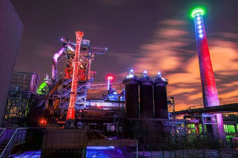 Night view of an illuminated industrial plant with colored lights and clouds in the sky,