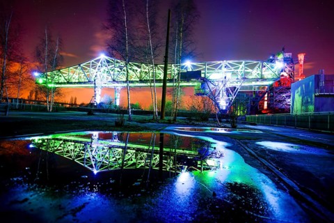 Illuminated bridge at night with blue and green lighting reflecting in a pool of water,