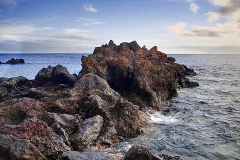 Rough rock formation jutting into the sea surrounded by waves under a slightly cloudy sky, Puerto