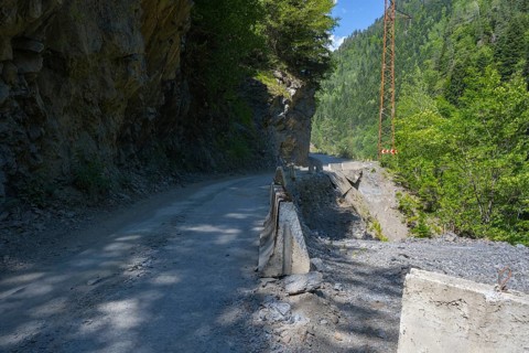 A narrow mountain road leads between rocky cliffs and green forest. Sun and shade areas alternate,