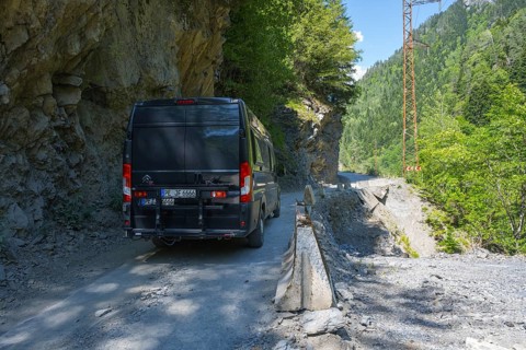 A black van drives on a narrow mountain road surrounded by rocks and thick vegetation, camper