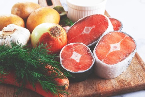 Potatoes, salmon, garlic, onion, and herbs are placed on a wooden cutting board. These ingredients
