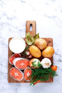Potatoes, salmon, garlic, onion, and herbs are placed on a wooden cutting board. These ingredients